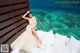 A woman in a wedding dress sitting on a bed by the ocean.